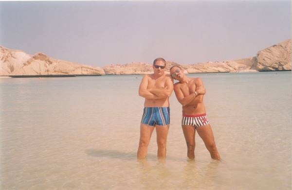 Two men in swim trunks hugging and laughing in clear water among white rocks