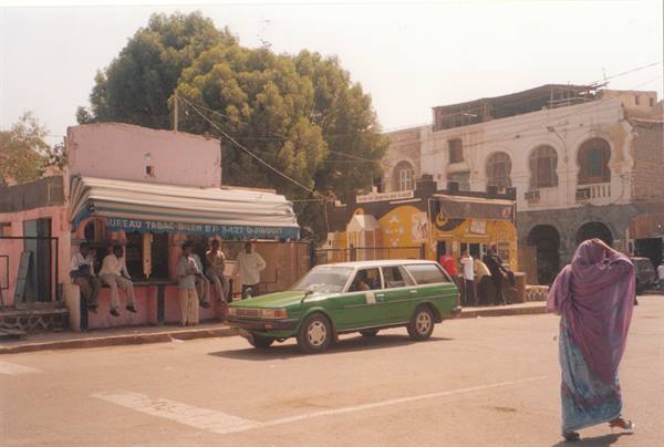 Woman in purple veil crossing the street, green car parked outside a café