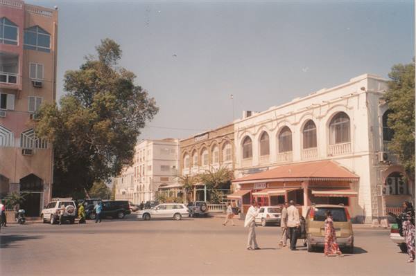 Pedestrians and cars on a square with arched colonial buildings, bright sunlight