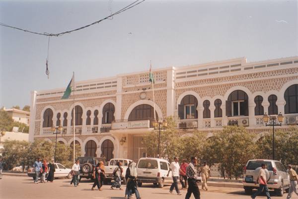 Pedestrians and cars in front of a white arched building, flags flying