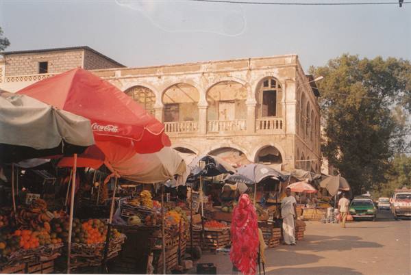 Open-air market with umbrellas, fruit crates, woman in pink dress before an arched colonial building