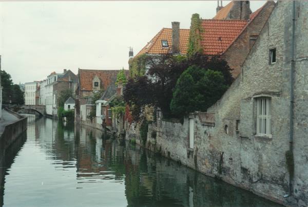 Brick and stone houses reflect in a calm Bruges canal under grey sky