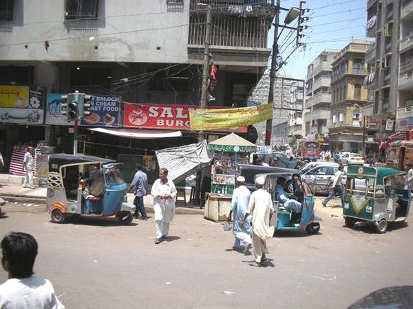 Green and blue rickshaws cross pedestrians in shalwar kameez on busy street