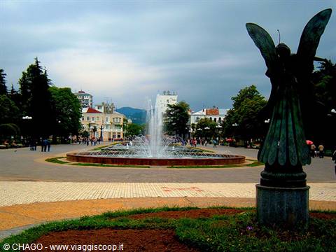 Bronze angel statue in front of a fountain in a tree-lined square