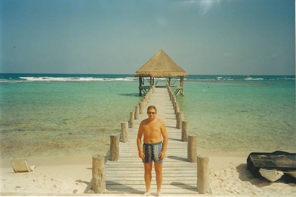 Man in swim trunks on wooden pier leading to thatched palapa over turquoise sea
