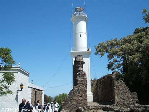 White lighthouse atop old stone ruins, tourists under tree, blue sky