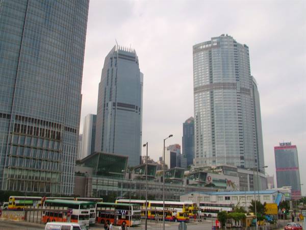 Glass skyscrapers in Hong Kong, double-decker buses waiting below under cloudy sky