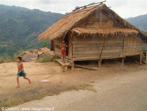 Children run past a wooden stilt house with thatched roof