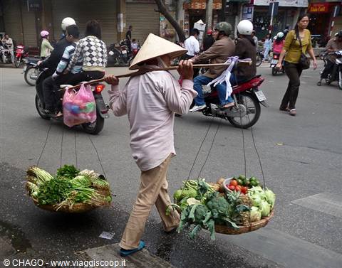 Street vendor carrying vegetable baskets on a pole among Hanoi motorbikes
