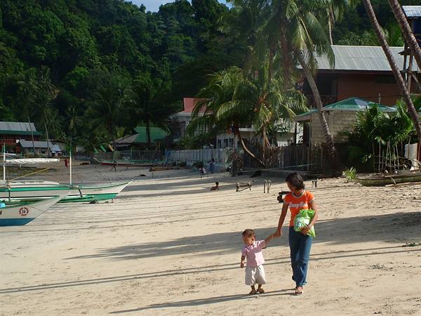 Mother and toddler walk on the beach past green boats and palms