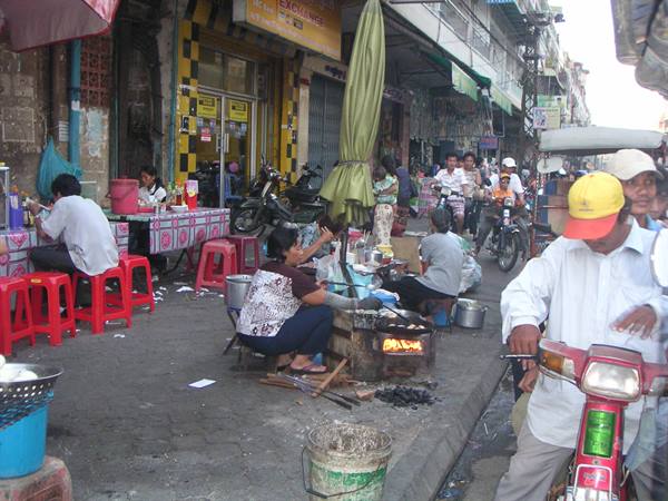Busy street market, woman cooking on charcoal grill, scooters and green umbrellas