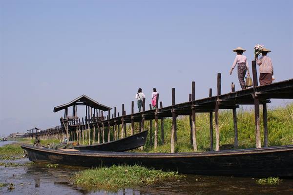 Wooden footbridge over Inle Lake, women carrying baskets walk, boats along the water