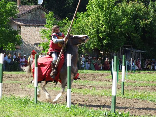 Armored knight with lance gallops between quintain poles, crowd in medieval costume