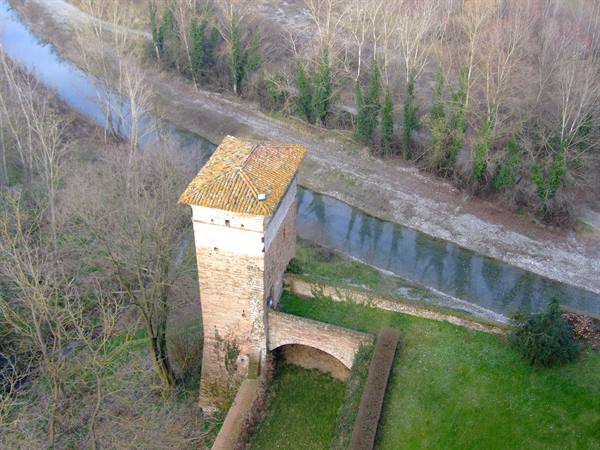 Brick tower with tiled roof beside a river among bare trees