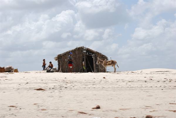 Wooden hut on sandy dunes, people and donkey under white clouds