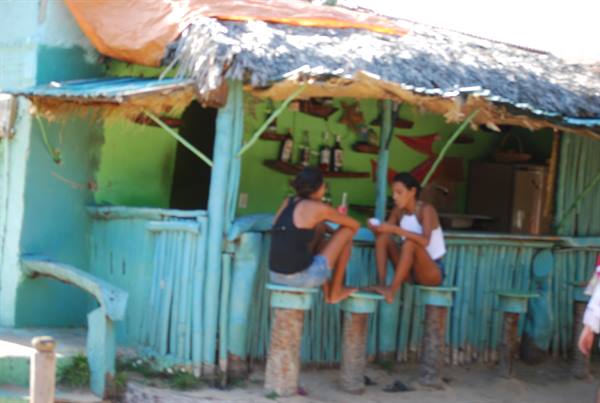 Two women sitting at the wooden counter of a thatched-roof beach bar