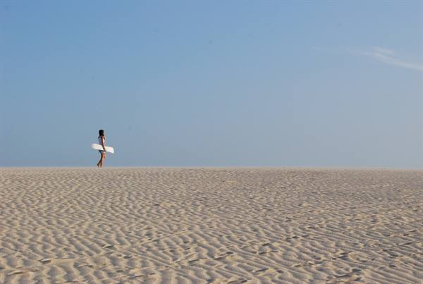 Woman carrying a surfboard walks across rippled sand, clear blue sky