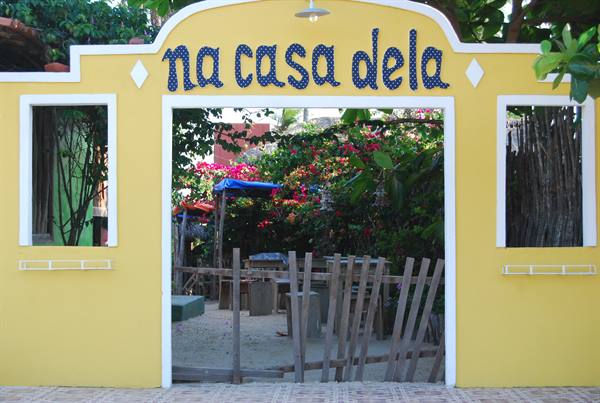 Yellow entrance with blue "na casa dela" sign, wooden gate and flowers