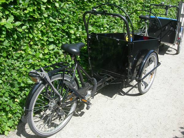 Black cargo bike parked beside green hedge on gravel path