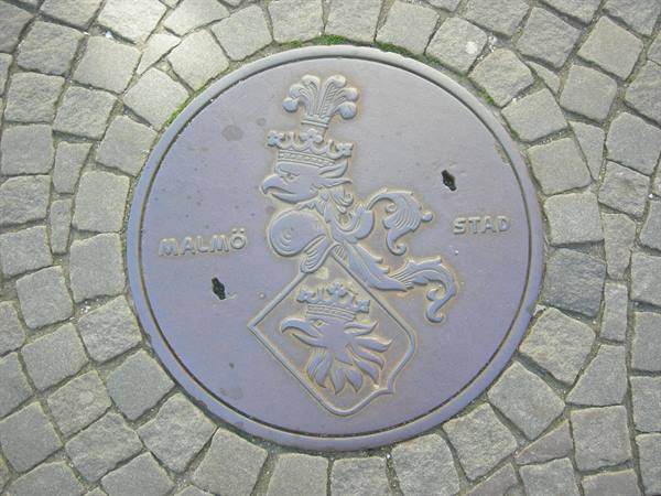 Metal manhole cover with griffin crest, Malmö Stad, set in cobblestones