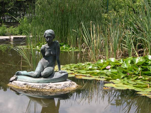 Bronze statue of seated woman on rock in pond among water lilies and reeds