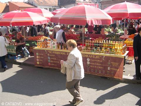 Woman walks past honey and fruit stall under red umbrellas at market