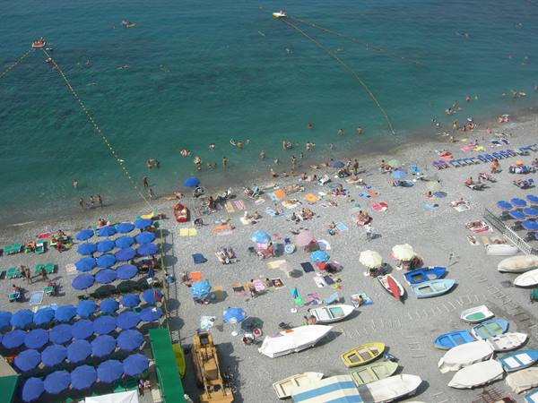 Pebble beach with blue umbrellas and boats ashore, swimmers in turquoise water