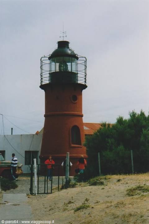 Red brick lighthouse with black lantern, visitors at the gate, overcast sky