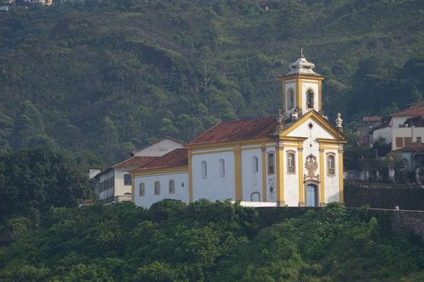 White and yellow colonial church with bell tower among houses and greenery in Ouro Preto