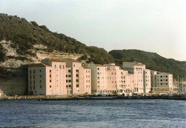 Pastel waterfront buildings below green hillside, boats moored along the quay