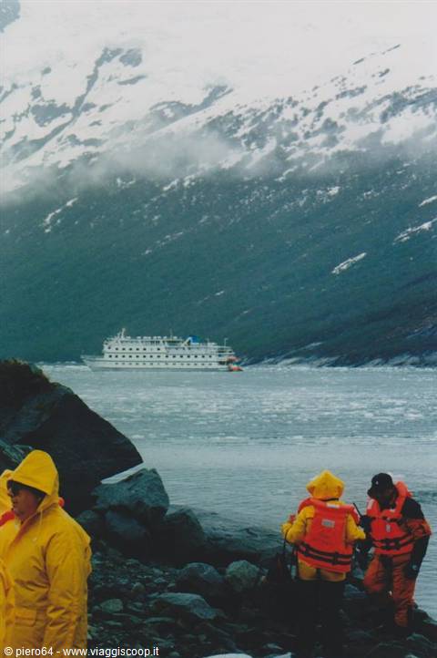 Tourists in yellow jackets on rocky shore, white ship among snowy mountains and ice