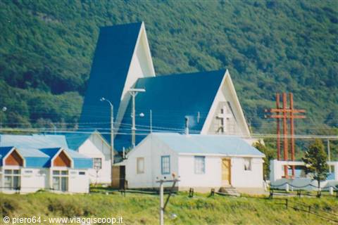 Blue-roofed church with tall spire and red cross below green mountains