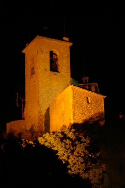 Stone bell tower lit golden at night, seen behind dark trees