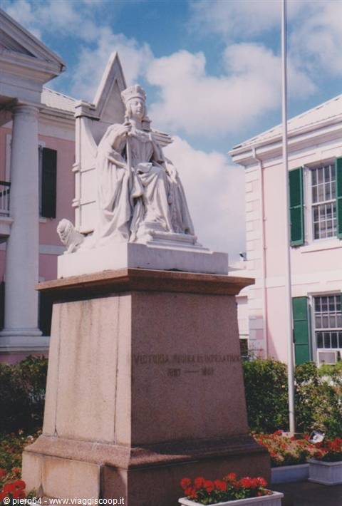 Marble statue of Queen Victoria seated on throne, in front of pink colonial building