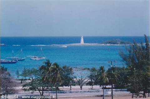 White lighthouse on islet, turquoise sea with boats, palm trees in foreground