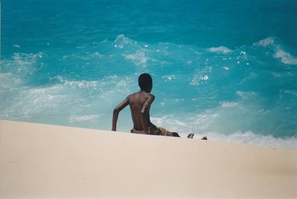 Boy sitting on white sand in front of turquoise waves