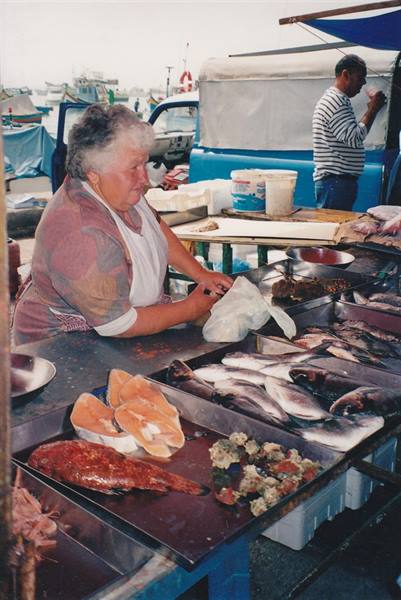Fish vendor at harbor market stall, metal trays with fresh fish, boats behind