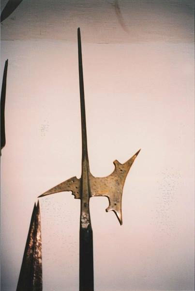 Metal halberd with angular blade hung on a pale wall beside a spear tip