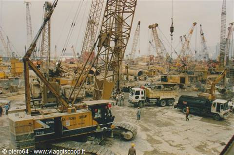 Construction yard with many yellow crawler cranes, trucks and workers under grey sky