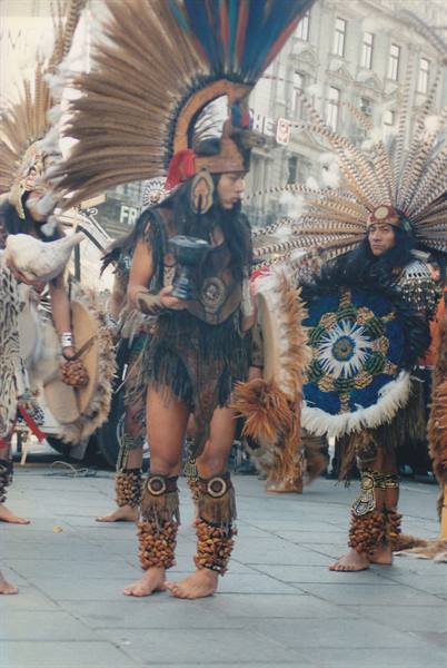 Aztec dancers in feather headdresses and shell anklets perform on a city street