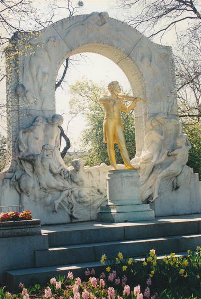 Golden Johann Strauss violin statue under marble arch, flowers in park