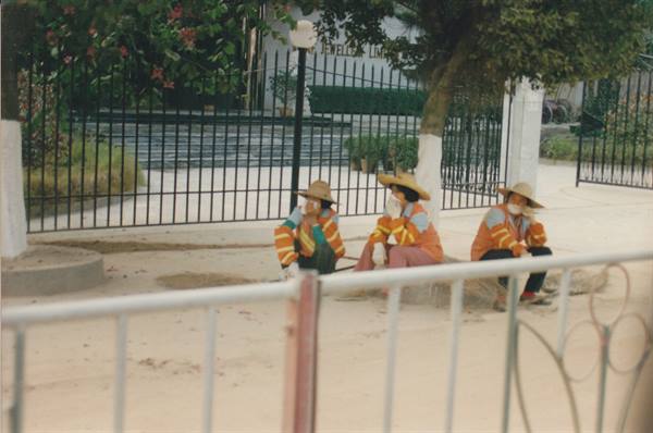 Three workers in straw hats and orange jackets rest beside a black fence