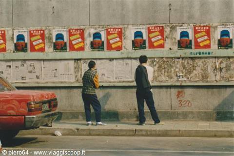 Two men read posters pasted on a city wall, red car parked nearby