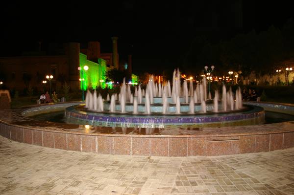 Illuminated circular fountain at night, water jets over stone, green-lit buildings behind