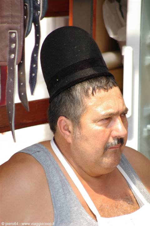 Moustached man wearing a tall black hat, in a shop with leather straps hanging