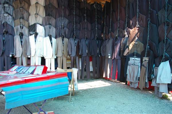 Open-air tailoring stall: suits hanging from chains, table with colorful fabrics