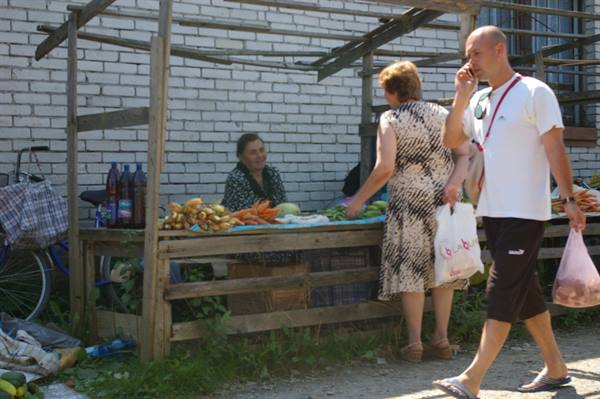 Vendor at wooden produce stall, customers with bags walking past