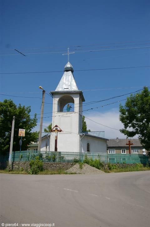 Small white church with metal-domed bell tower and cross, behind green picket fence