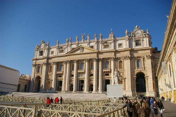 St. Peter's Basilica facade with columns and statues under blue sky, tourists walking