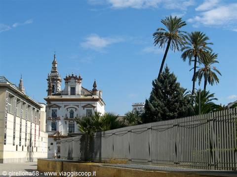 White baroque church with bell tower, palm trees and fence under blue sky